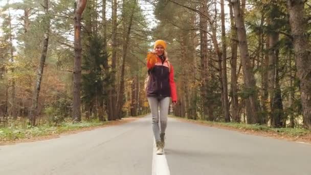 Una giovane ragazza magra con un cappello giallo e uno zaino vintage cammina lungo una strada asfaltata nella foresta di conifere giallo autunno. Vista frontale — Video Stock