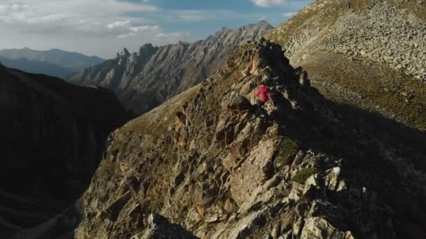 Un joven con barba, un montañero con gorra y gafas de sol, sube a una cresta rocosa en lo alto de las montañas. Parkour en las montañas. Opinión Arial — Vídeos de Stock