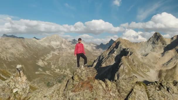 Bir sakallı bir genç hipster adam bir tırmanıcı bir şapka ve güneş gözlüğü dağlarda yüksek kayalık bir sırt üzerinde duran ve etrafa. Parkour dağlarda. Airview görünümü. Hareketinden — Stok video