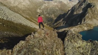 Bir sakallı bir genç hipster adam bir tırmanıcı bir şapka ve güneş gözlüğü dağlarda yüksek kayalık bir sırt üzerinde duran ve etrafa. Parkour dağlarda. Airview görünümü. Hareketi çevresinde