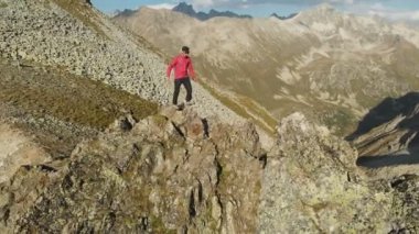 Bir sakallı bir genç hipster adam bir tırmanıcı bir şapka ve güneş gözlüğü dağlarda yüksek kayalık bir sırt üzerinde duran ve etrafa. Parkour dağlarda. Airview görünümü. Hareketi çevresinde