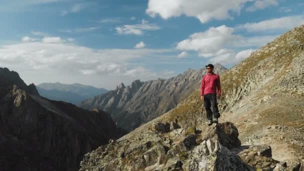Um jovem hipster com uma barba um alpinista em um boné e óculos de sol está de pé em um cume rochoso alto nas montanhas e olhando ao redor. Parkour nas montanhas. Vista aérea. Circulação — Vídeo de Stock