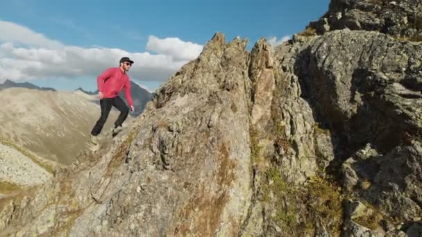 Молодой парень с бородой, альпинист в кепке и солнцезащитных очках, взбирается на скалистый хребет высоко в горах. Паркур в горах. Arial view — стоковое видео
