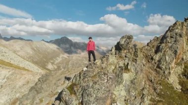 Bir sakallı bir genç hipster adam bir tırmanıcı bir şapka ve güneş gözlüğü dağlarda yüksek kayalık bir sırt üzerinde duran ve etrafa. Parkour dağlarda. Airview görünümü. Hareketinden