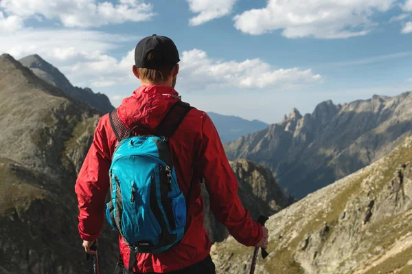 Vousatý muž v sluneční brýle a čepici s batohem stojí na vrcholu skály a dívá do vysoké skalnaté údolí v horách. Koncepce cestovního ruchu a snadný trekking v horách venkovní — Stock fotografie