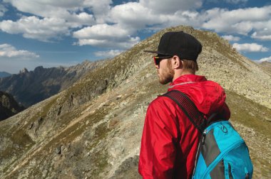 Sakallı bir adam güneş gözlüğü ve bir sırt çantası ile bir kap içinde bir kaya üzerine anlamına gelir ve bir kayalık vadide yüksek dağların içine görünüyor. Turizm ve kolay açık dağlarda trekking kavramı