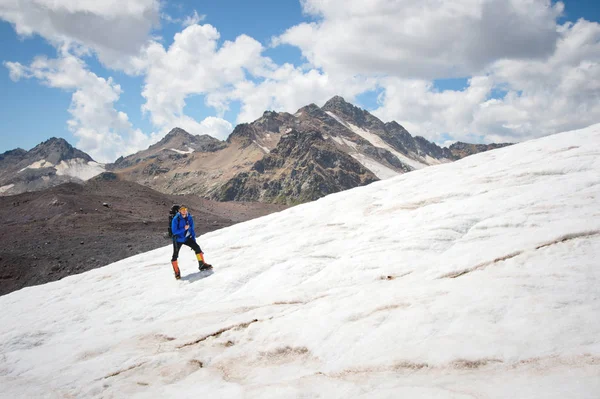 Reiziger in een pet en zonnebril met een rugzak op zijn schouders in de besneeuwde bergen op de gletsjer tegen de lucht en de wolken. Reiziger in een natuurlijke omgeving — Stockfoto