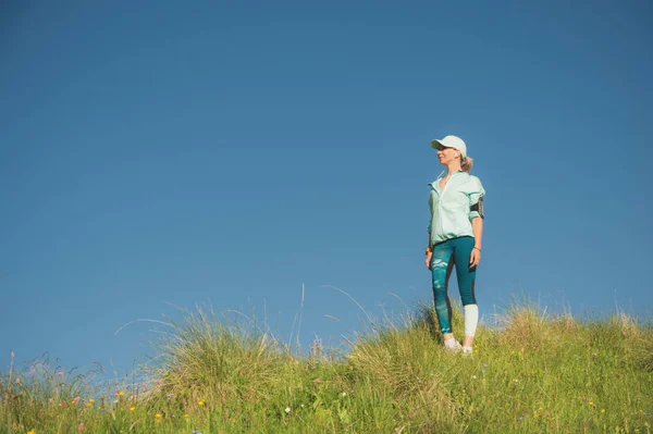 Fitness runner woman listening to music on the nature. Portrait of beautiful girl wearing earphones earbuds and running cap. against the background of a green field and blue sky — Stock Photo, Image