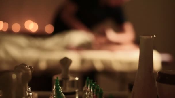 Un grande massaggiatore fa un massaggio per una giovane ragazza sdraiata sul divano a faccia in giù in un ufficio buio con candele sullo sfondo. Vista da dietro gli accessori per il massaggio sfocare chiave scura — Video Stock