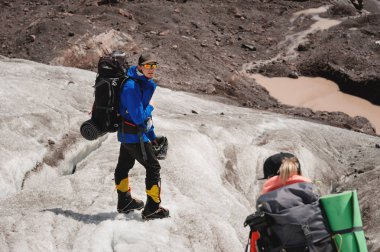 İki turist, bir erkek ve bir kadın sırt çantaları ve krampon kendi ayakları üzerinde buzul dağ Elbrus ve gökyüzü ve bulutlar arka planı boyunca yürümek. Arkadan görünüm