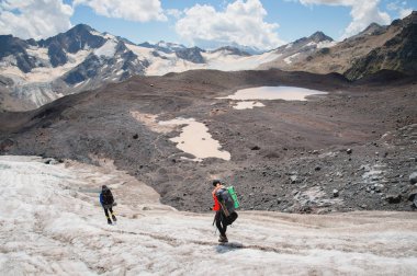 İki turist, bir erkek ve bir kadın sırt çantaları ve krampon kendi ayakları üzerinde buzul dağ Elbrus ve gökyüzü ve bulutlar arka planı boyunca yürümek. Arkadan görünüm