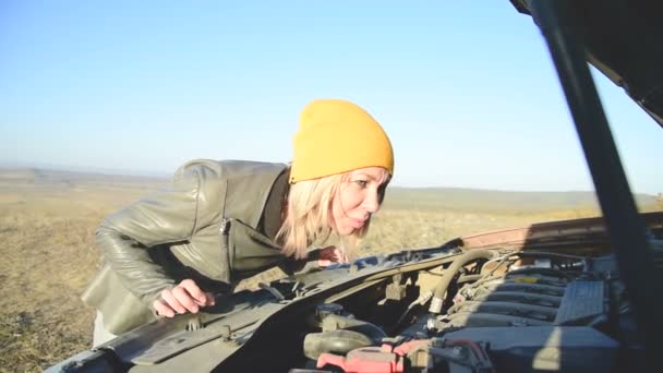 La rubia en la gorra mira debajo de la capucha del coche está buscando una falla pero no sabe exactamente dónde buscar — Vídeo de stock