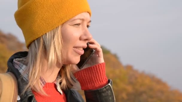 Närbild en attraktiv kaukasiska tjej i en gul hatt talar på sin mobiltelefon utomhus på hösten. Leende flicka i kommunikation — Stockvideo