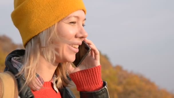 Close-up Una atractiva chica caucásica con un sombrero amarillo está hablando en su teléfono celular al aire libre en otoño. Chica sonriente en la comunicación — Vídeos de Stock