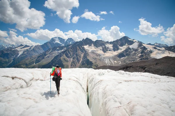 Женщина-альпинистка наслаждается красотой ледника, прогуливаясь по леднику в кошки и солнцезащитных очках. На фоне высоких гор Кавказа небо и облака — стоковое фото