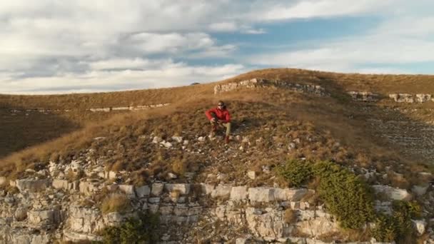 Una vista aérea de un macho hipster barbudo con un control remoto de un dron camina a lo largo del borde de una meseta alta cerca de un acantilado al atardecer. El tipo está sentado en el acantilado. — Vídeos de Stock