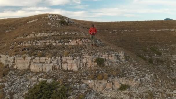 Una vista aerea di un maschio hipster barbuto con un telecomando da un drone cammina lungo il bordo di un altopiano vicino a una scogliera al tramonto. Il tizio è sulla scogliera. — Video Stock