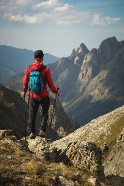 Sakallı bir adam güneş gözlüğü ve bir sırt çantası ile bir kap içinde bir kaya üzerine anlamına gelir ve bir kayalık vadide yüksek dağların içine görünüyor. Turizm ve kolay açık dağlarda trekking kavramı