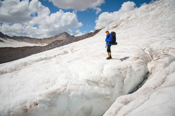 Путешественник в кепке и солнечных очках с рюкзаком на плечах в заснеженных горах на леднике против неба и облаков. Путешественник в естественной среде — стоковое фото