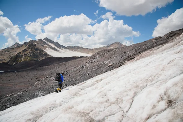 Reiziger in een pet en zonnebril met een rugzak op zijn schouders in de besneeuwde bergen op de gletsjer tegen de lucht en de wolken. Reiziger in een natuurlijke omgeving — Stockfoto