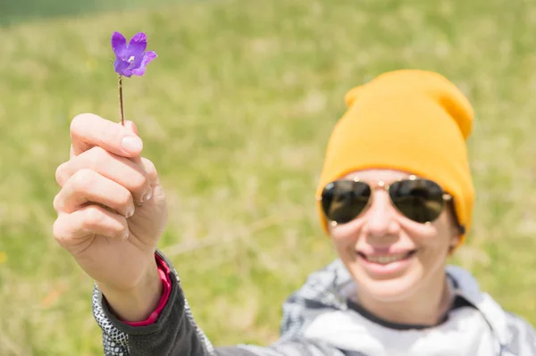 O retrato de uma menina alegre feliz em um chapéu e óculos de sol dá-lhe uma flor de campo contra um fundo de campos verdes — Fotografia de Stock