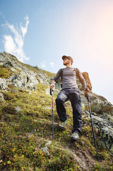 Vousatý muž v sluneční brýle a čepici s batohem stojí na vrcholu skály a dívá do vysoké skalnaté údolí v horách. Koncepce cestovního ruchu a snadný trekking v horách venkovní — Stock fotografie