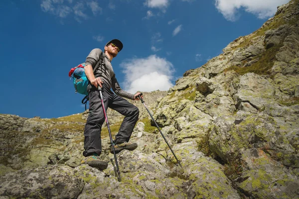 Γενειοφόρος άνδρας με γυαλιά ηλίου και ένα καπάκι με ένα σακίδιο στέκεται πάνω σε ένα βράχο και μοιάζει σε μια βραχώδη κοιλάδα ψηλά στα βουνά. Η έννοια του τουρισμού και εύκολη πεζοπορία στα βουνά εξωτερική — Φωτογραφία Αρχείου