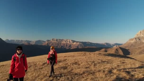 Vista aérea de un viajero de dos chicas con mochilas y cámaras paseando por las colinas entre las rocas épicas de las montañas. Chicas fotógrafas con sus cámaras al atardecer — Vídeos de Stock