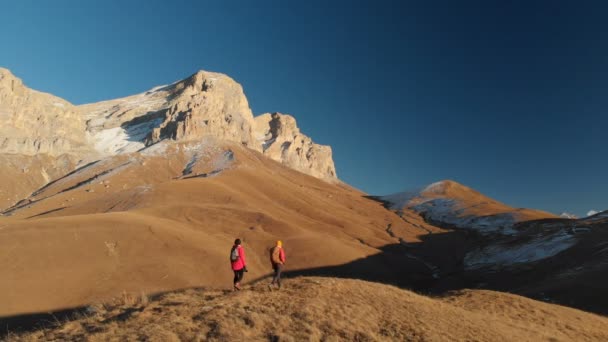 Vista aérea de un viajero de dos chicas con mochilas y cámaras paseando por las colinas entre las rocas épicas de las montañas. Chicas fotógrafas con sus cámaras al atardecer — Vídeos de Stock