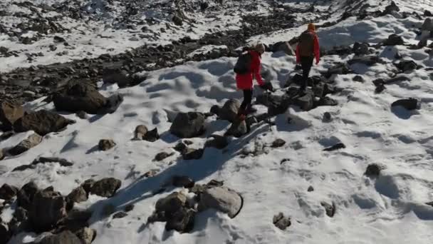 Vista aérea de dos chicas viajeras con mochilas y cámaras que atraviesan la nieve y las piedras hasta el glaciar entre las rocas épicas de las montañas. Chicas fotógrafas con sus cámaras al atardecer — Vídeos de Stock