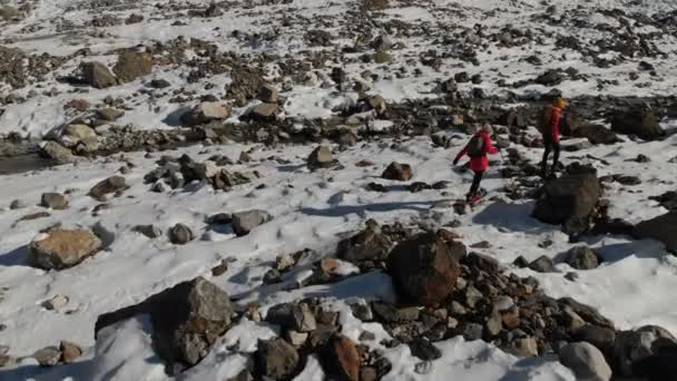 Letecký pohled na dvě dívky cestovatel s batohy a fotoaparáty projít sněhu a kamení na ledovec mezi impozantní skály v horách. Holky fotografů s jejich fotoaparáty při západu slunce — Stock video