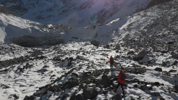 Luftaufnahme von zwei Mädchen Reisende mit Rucksäcken und Kameras gehen durch den Schnee und Steine auf den Gletscher zwischen den epischen Felsen in den Bergen. Fotografinnen mit ihren Kameras bei Sonnenuntergang — Stockvideo