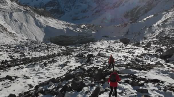 Vista aérea de duas meninas viajante com mochilas e câmeras atravessam a neve e pedras para a geleira entre as rochas épicas nas montanhas. Fotógrafos meninas com suas câmeras ao pôr do sol — Vídeo de Stock