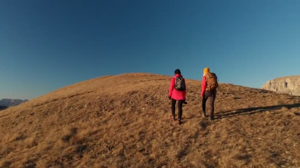 Vista aérea de dos chicas viajeras con mochilas y cámaras suben la colina entre las rocas épicas de las montañas. Chicas fotógrafas con sus cámaras al atardecer — Vídeo de stock