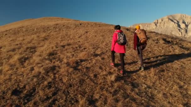 Vista aérea de dos chicas viajeras con mochilas y cámaras suben la colina entre las rocas épicas de las montañas. Chicas fotógrafas con sus cámaras al atardecer — Vídeos de Stock