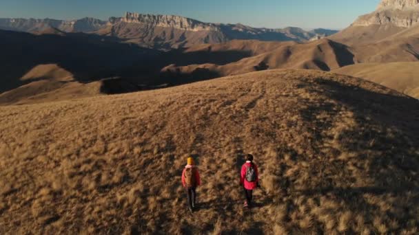 Vista aérea de un viajero de dos chicas con mochilas y cámaras paseando por las colinas entre las rocas épicas de las montañas. Chicas fotógrafas con sus cámaras al atardecer — Vídeo de stock