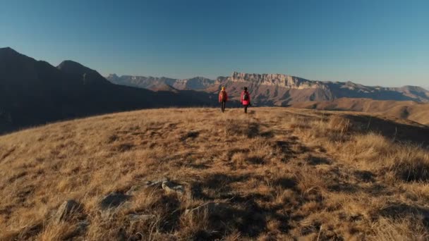 Vista aérea de un viajero de dos chicas con mochilas y cámaras paseando por las colinas entre las rocas épicas de las montañas. Chicas fotógrafas con sus cámaras al atardecer — Vídeos de Stock