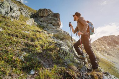 Sakallı bir adam güneş gözlüğü ve bir sırt çantası ile bir kap içinde bir kaya üzerine anlamına gelir ve bir kayalık vadide yüksek dağların içine görünüyor. Turizm ve kolay açık dağlarda trekking kavramı