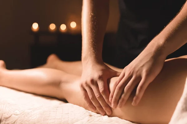 Voor een vrouw doet een masseur in een donkere kamer een hip massage met olie. Spa procedures massage vrouwen — Stockfoto