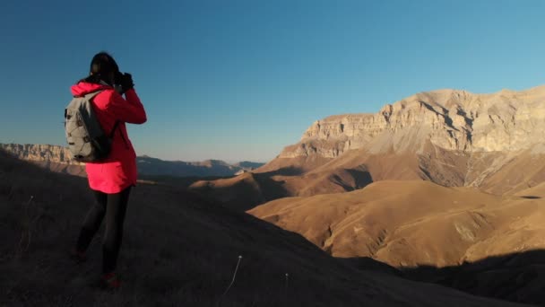 Flygfoto över en flicka resenär med en ryggsäck och en kamera tar bilder i bergen vid solnedgången. Flicka fotograf från baksidan. Kameran passerar förbi och flyger till bergen — Stockvideo