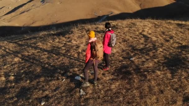 Vista aérea de un viajero de dos chicas con mochilas y cámaras paseando por las colinas entre las rocas épicas de las montañas. Chicas fotógrafas con sus cámaras al atardecer — Vídeos de Stock