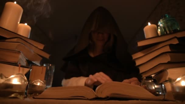 Medium close-up girl magician in a hood in a dark room by candlelight and looking for a spell turning over a book. Low key. Mystic Small DOF — Stock Video