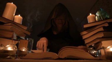 Mum ışığı ve seyir için bir kitap üzerinde bir büyü tarafından karanlık bir odada bir başlıklı orta yakın çekim kız sihirbaz. Düşük anahtar. Mistik küçük Dof