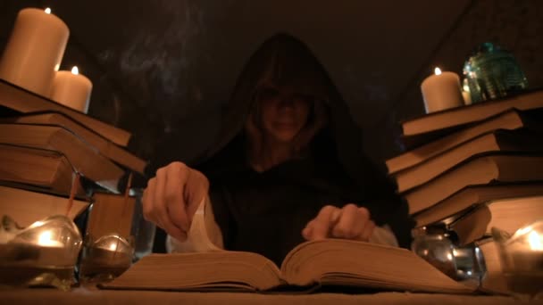 Μέτρια γκρο πλαν κορίτσι μάγος με κουκούλα σε ένα σκοτεινό δωμάτιο από το φως των κεριών και ψάχνει για ένα ξόρκι στροφή πάνω από ένα βιβλίο. Χαμηλών τόνων. Μυστικιστής μικρό Dof — Αρχείο Βίντεο