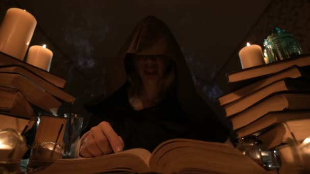 Střední detail dívka kouzelník v kapuci v temné místnosti při svíčkách a hledají kouzlo soustružení nad knihou. Nízký klíč. Mystic malé Dof — Stock video