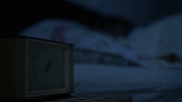 Primer plano de una chica en pijama acostada en la cama e incapaz de dormir mirando un reloj despertador. Enfoque variable de horas a cara con las chicas. El efecto de la noche americana. Noche de imitación de luz fría de llave baja — Vídeo de stock