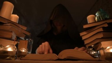 Mum ışığı ve seyir için bir kitap üzerinde bir büyü tarafından karanlık bir odada bir başlıklı orta yakın çekim kız sihirbaz. Düşük anahtar. Mistik küçük Dof