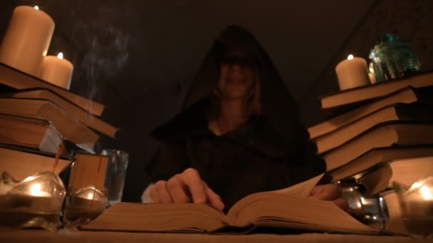 Średnich szczegół dziewczyna Mag w kaptur w ciemnym pomieszczeniu przy świecach i szukam zaklęcie przewracając książkę. Niski klucz. Mystic mały Dof — Wideo stockowe