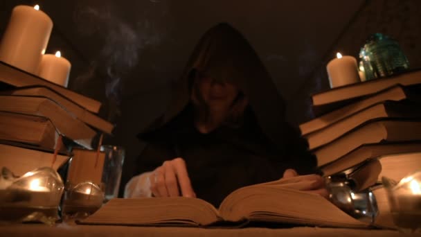 Média menina de close-up mágico em um capuz em um quarto escuro à luz de velas e à procura de um feitiço virar um livro. Chave baixa. Místico pequeno DOF — Vídeo de Stock
