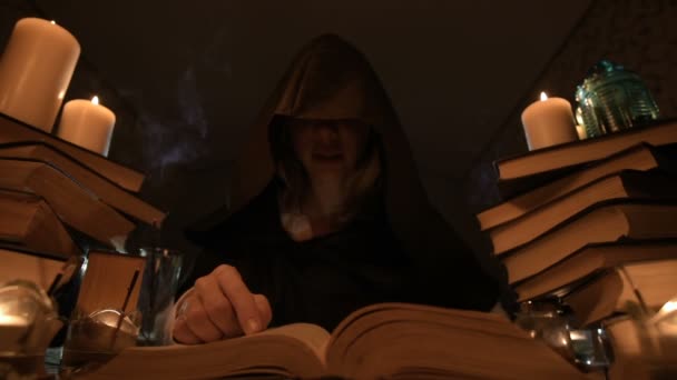 Medium close-up girl magician in a hood in a dark room by candlelight and looking for a spell turning over a book. Low key. Mystic Small DOF — Stock Video